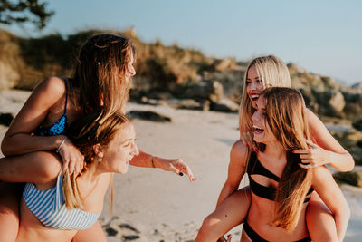 Cheerful millennial girlfriends in swimwear laughing happily while giving piggyback ride to each other during summer vacation on sandy beach