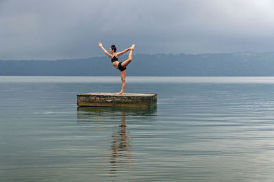 Woman in king dancer pose doing yoga on floating platform amidst lake against sky