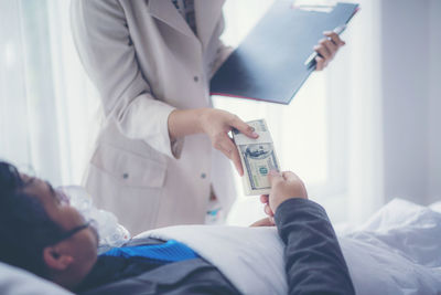 Midsection of businesswoman giving money to colleague lying on bed in hospital