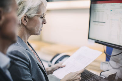 Businesswoman reading document while standing in front of desktop computer with colleague at industry