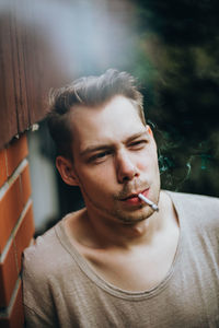 Young man smoking cigarette while leaning on brick wall