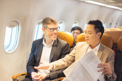 During a flight, a businessman in formal attire and glasses works with a friend near the window