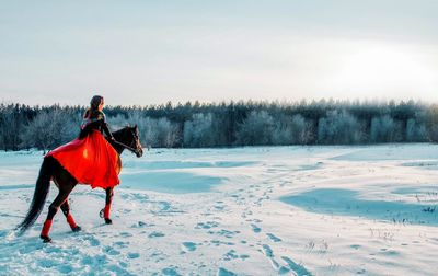 Rear view of woman riding horse on snow covered field against sky