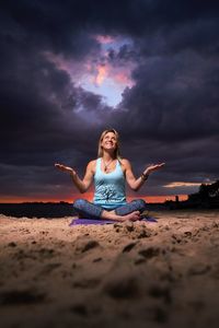 Full length of smiling woman looking up while sitting at beach against storm clouds