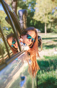 Portrait of happy young woman peeking through car window in forest