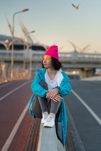 Thoughtful female hipster resting sitting enjoying sunset and warm weather on curbstone near road.