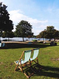 Chairs on grass by lake against sky