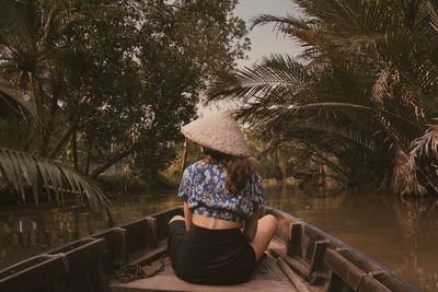 Rear view of woman wearing asian style conical hat sitting on rowboat in lake