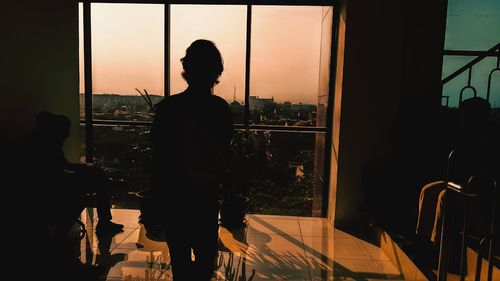 Silhouette man standing by window against sky during sunset