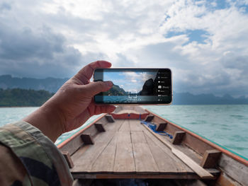 Cropped hand of man photographing sea with mobile phone against cloudy sky