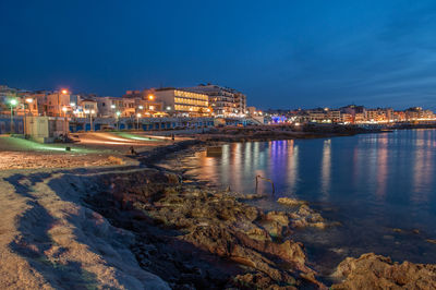 Illuminated buildings by sea against sky at night