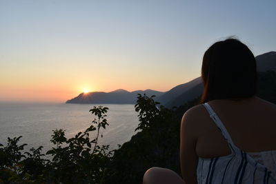 Rear view of woman looking at sea while sitting on mountain during sunset