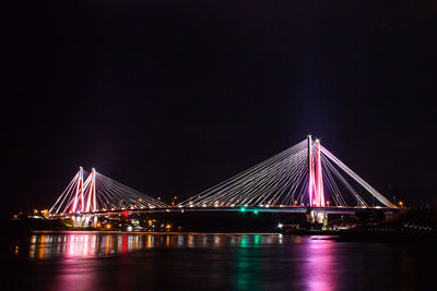 Illuminated suspension bridge over river against sky at night