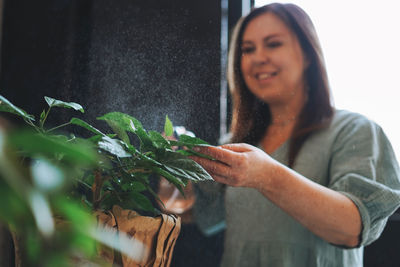 Adult middle aged brunette smiling woman sprays house plants by water in kitchen at home