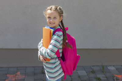 A cute schoolgirl with a knapsack behind her back smiles and holds colorful notebooks 