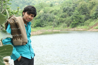 Young man smiling while standing by lake against trees