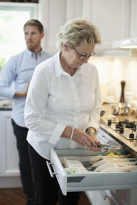Senior woman arranging cutlery with son standing in background at kitchen