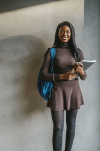 Full length portrait of smiling young woman holding book standing with backpack against gray wall