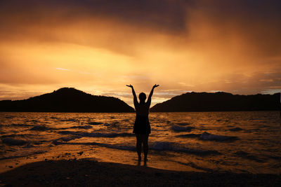 Rear view of silhouette woman with arms raised standing at beach during sunset