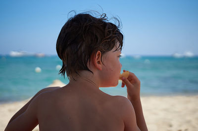 Rear view of shirtless boy eating food at beach