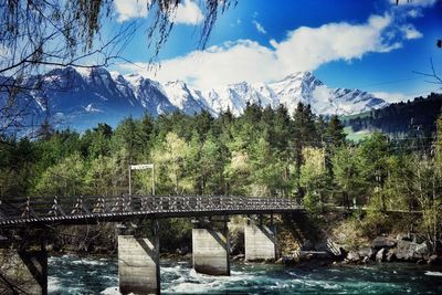 Bridge over river against trees and snowcapped mountains