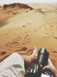 Low section of man holding camera sitting on sand at desert