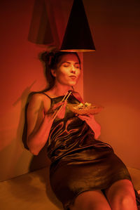 Young woman looking away while sitting against orange wall