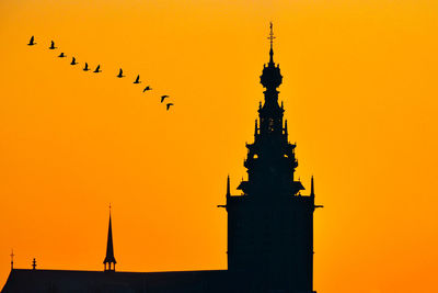 Silhouette birds flying by building against orange sky during sunset