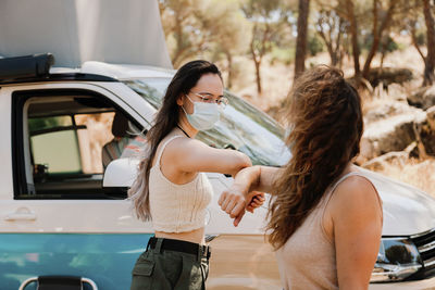 Friendly women in protective masks standing near van in forest and bumping elbows while greeting each other during coronavirus epidemic