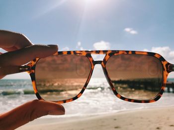 Close-up of human hand holding sunglasses at beach against sky during sunny day