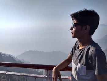 Young man standing on railing by sea against sky