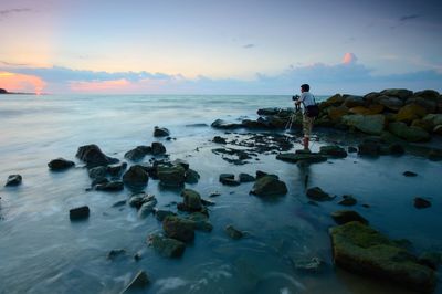 People standing on rocks at beach against sky during sunset