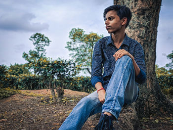 Young man looking away while sitting on tree trunk