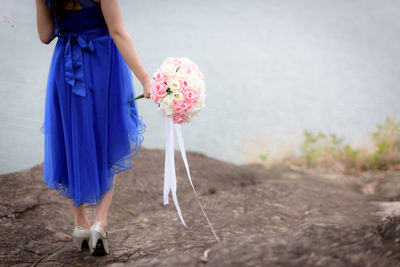 Low section of bride with bouquet standing on rock formation by lake