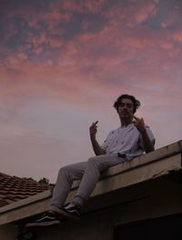 Young man sitting on roof against sky during sunset