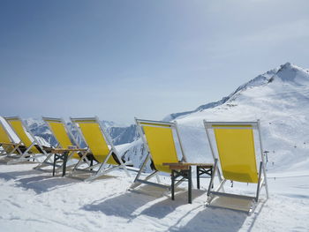 Empty yellow deck chairs by snow covered mountain against clear sky