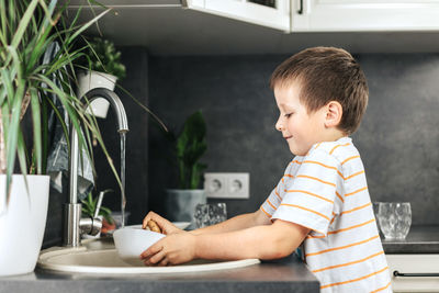 Cute boy washes dishes with a wooden brush with natural bristles in the kitchen