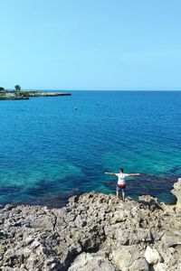 High angle view of man with arms outstretched standing at rocky beach against clear blue sky