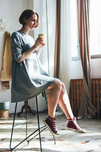 Young woman drinking coffee while sitting on chair at home