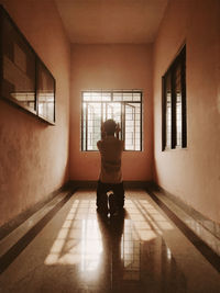 Rear view of man kneeling while praying on tiled floor by window in corridor