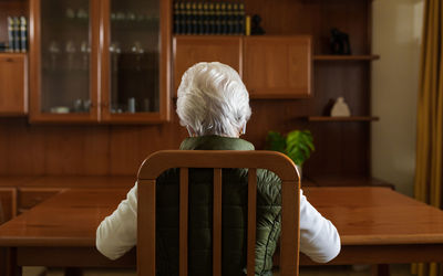 Back view of anonymous elderly female patient in tws earbuds speaking with doctor on tablet during video call at home