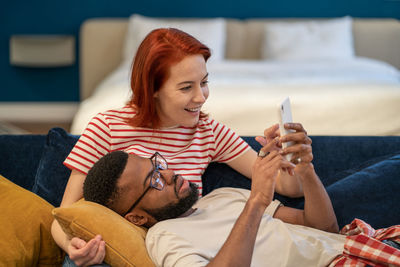 Multi ethnic happy couple man and woman look at phone lying on couch in living room.