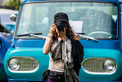 Young woman holding camera while standing against car