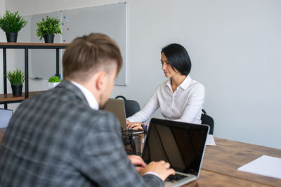 A man in a jacket is working at a laptop next to a woman in the office