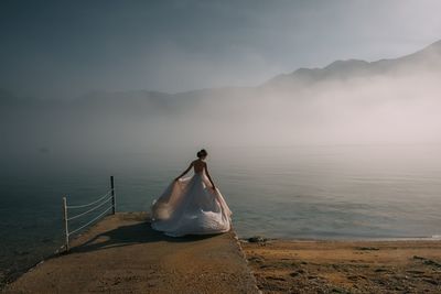 Rear view of bride standing at lakeshore against sky