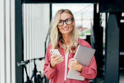 Confident businesswoman uses phone while standing with laptop at exit from office building.