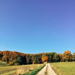 Road passing through field against clear blue sky