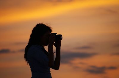 Side view of silhouette woman photographing against orange sky