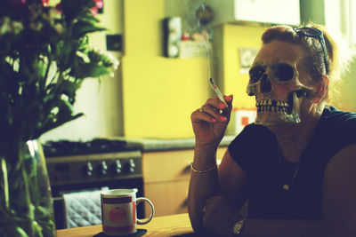 Woman wearing mask smoking cigarette while sitting by table at home