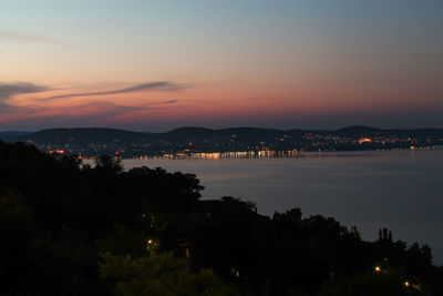 Illuminated city by sea against sky at sunset